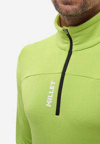 Grüner Fleece-Oberteil mit Reißverschluss und hohem Kragen, mit schwarzem Reißverschluss und Akzenten. "Millet"-Logo in Weiß auf der Vorderseite gedruckt. Glatte Textur.