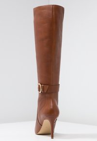 Botte haute en cuir marron, à la coupe ajustée, bout pointu et talon aiguille. Présente un accent de boucle près de la cheville.