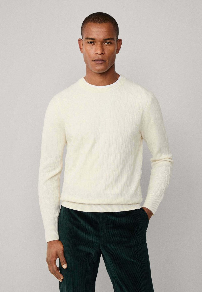 Suéter de punto trenzado en crema con cuello redondo y puños acanalados, combinado con pantalones de pana verde oscuro. Diseño simple y texturizado.
