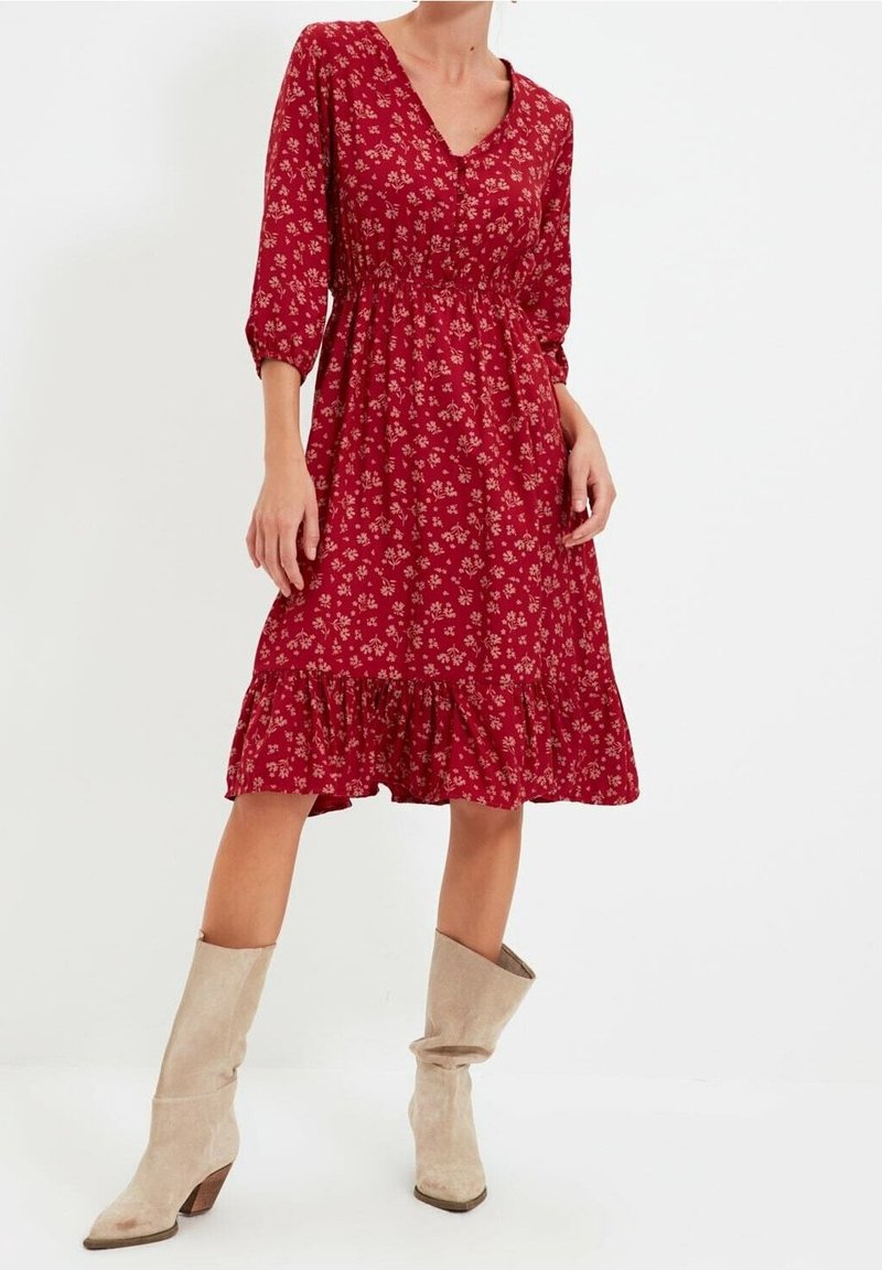 Femme portant une robe midi rouge à fleurs avec des manches trois-quarts et des bottes beiges en daim décontractées mi-mollet, debout sur un fond blanc.