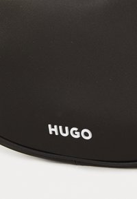 Černá látková taška s texturovaným povrchem. Na přední straně se nachází bílé logo "HUGO". Zaoblený tvar s zipem na vrchu.