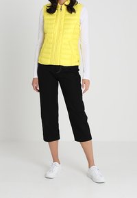 Gelbe, gesteppte Pufferweste mit hohem Kragen, Reißverschluss und kombiniert mit schwarzen, cropped Hosen sowie weißen Sneakers. Minimalistisches Design und lässige Passform.