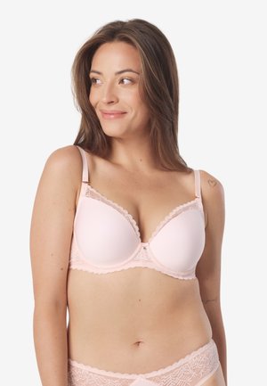 Kobieta z długimi brązowymi włosami, ubrana w jasnoróżowy koronkowy biustonosz i pasujące majtki, uśmiechająca się i patrząca w lewo.