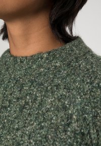 Grön texturerad stickad tröja med hög hals. Materialet verkar mjukt med ett varierat mönster. Tyget har en något fluffig textur.