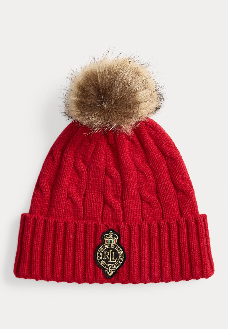 Gorro de malha vermelho com padrão de cabos, apresentando um pom-pom de pelo castanho no topo e um emblema preto e dourado na bainha dobrada canelada.
