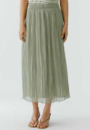 Femme portant une jupe midi plissée vert clair avec une taille élastique et des sandales marron à brides, debout devant un fond uni.