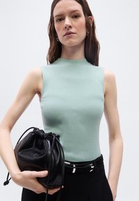 Γυναίκα με αμάνικο ανοιχτό πράσινο τοπ που κρατάει μαύρη δερμάτινη τσάντα με κορδόνι, σε λευκό φόντο.