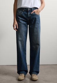 Jeans blu a gamba larga in denim, con vita alta, chiusura con bottoni e tasche frontali. Abbinati a scarpe beige.