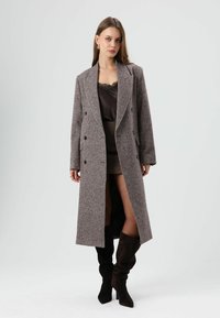 Dubbelgeknoopte, lange grijsbruine jas met textuurrijk materiaal, zijzakken en op maat gemaakte revers. Draagt over een donker satijnen jurk en kniehoge laarzen.