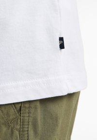 Parte inferior de una camiseta blanca de algodón con una etiqueta de marca tejida; combinada con pantalones de tela verde oliva que muestran detalles de costura en los bolsillos y texturas.