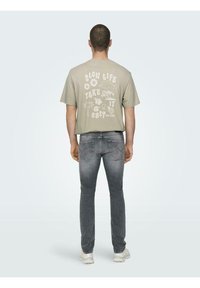 Un tricou scurt, de culoare bej deschis, cu mâneci scurte, are un text grafic alb și ilustrații pe spate, fiind asortat cu blugi gri și adidași albi.