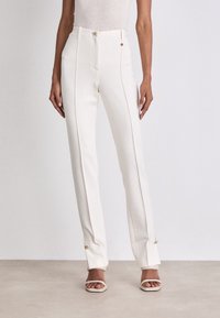 Pantalon blanc sur mesure avec une taille cintrée, des plis à l'avant et des accents en métal doré. Tissu lisse et jambes évasées qui valorisent la silhouette.