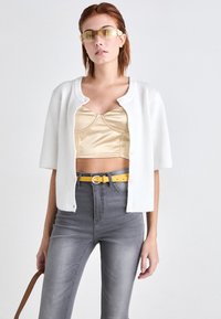 White ribbed cardigan with short sleeves, over a beige satin cropped top. Gray high-waisted jeans with a yellow belt. Yellow-tinted sunglasses.