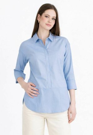 Frau mit langen braunen Haaren, die ein hellblaues Hemd mit Knöpfen und Dreiviertelärmeln sowie cremefarbene Hosen trägt, steht mit einer Hand in der Hüfte.