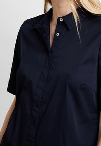 Donna che indossa una camicia a maniche corte blu scuro con colletto su uno sfondo chiaro e semplice.