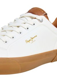 Witte sneaker met een glad ontwerp, een zool van geribbeld rubber en bruine leren accenten bij de hiel. Gouden logo aan de zijkant. Witte veters.