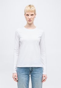 Ξανθός με κοντά μαλλιά που φοράει ασημένια σκουλαρίκια κρίκους, ένα απλό λευκό μακρυμάνικο πουκάμισο και ανοιχτό μπλε τζιν, στέκεται μπροστά σε λευκό φόντο.