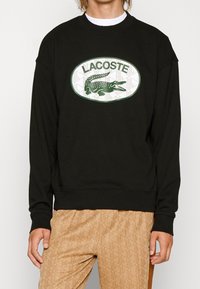 Sudadera negra con un gran logotipo ovalado verde de Lacoste que presenta un diseño de cocodrilo, con puños y dobladillo de canalé, fabricada en un tejido suave.