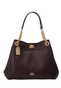 Brązowa skórzana torebka o fakturowanym wykończeniu, z złotymi łańcuszkowymi ramiączkami, okrągłym magnetycznym zapięciem i małą zawieszką. Logo COACH wytłoczone na dole.