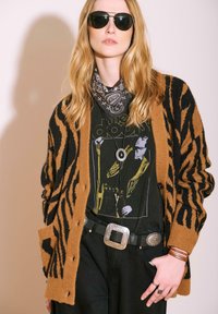 Femme portant des lunettes de soleil noires, un cardigan à rayures tigre brun et noir, un t-shirt graphique de groupe, un pantalon noir, une ceinture large et un foulard en bandana noir.