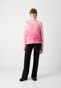 Een persoon die staat, gekleed in een roze gebreid trui, zwarte wijde broek met kleine studs, zwarte schoenen en een zilveren clutch vasthoudend.