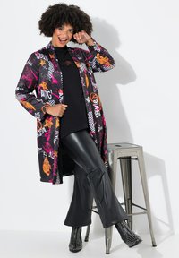 Abrigo largo y negro con patrones gráficos coloridos, combinado con una blusa negra y pantalones de campana negros brillantes. El calzado tiene tachuelas metálicas.