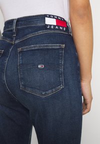 Vaqueros de mezclilla azul oscuro con forma ajustada, que cuentan con un bolsillo trasero y un parche de logo de Tommy Jeans en rojo, blanco y azul en la cintura.