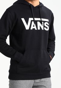 Persona che indossa una felpa nera Vans con tasca anteriore e stringhe, mani in tasca, abbinata a pantaloni grigi.