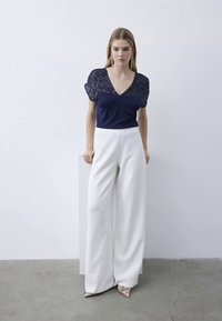 Top blu navy con spalle decorate da paillettes, scollatura a V; pantaloni bianchi a gamba larga; tessuto liscio; design minimalista; modella in posa contro uno sfondo chiaro.