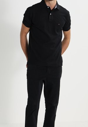 Mężczyzna stojący z rękami za plecami, ubrany w czarną krótka koszulkę polo z krótkim rękawem i czarne spodnie na jednolitym tle.