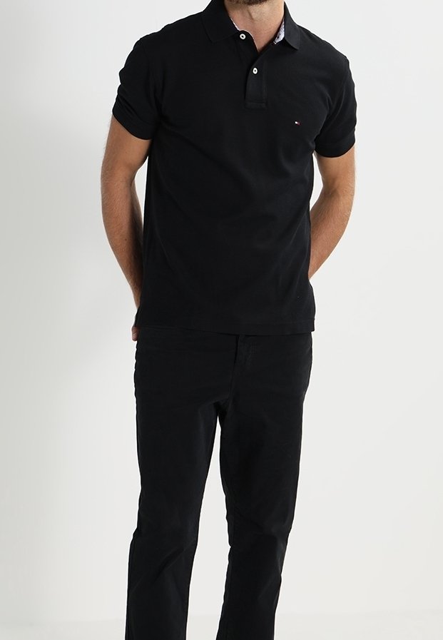 Homme debout, mains derrière le dos, portant un polo noir à manches courtes et un pantalon noir, sur un fond uni.