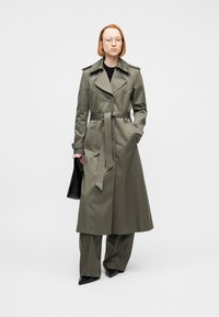 Femme aux cheveux rouges courts portant des lunettes, un long trench-coat olive, un pantalon à jambes larges, des chaussures noires pointues, tenant une pochette noire.