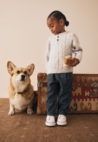 Een kind in een lichtgrijze kabelgebreide trui met knopen, een broek met patroon en witte sneakers staat naast een corgi bij een vintage koffer.