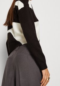 Svart och krämfärgad mönstrad tröja med strukturerad yta, kombinerad med grå byxor. Noterbara detaljer inkluderar en boxig passform och sidofickestyle.