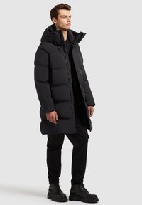 Abrigo acolchado negro con capucha, con cierre delantero, diseño acolchado y una gran aislación. Llevado con pantalones negros y botas de combate.