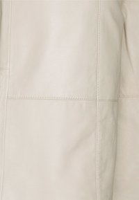 Tessuto in pelle di un leggero beige con una texture liscia, caratterizzato da cuciture pulite e un leggero lucido. Nessun motivo o hardware visibile.