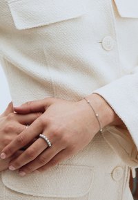 Hopeinen koruvalikoima sisältää pyöreän sormuksen, jossa on useita kirkkaita kiviä, ja herkkä rannekoru, jossa on pieniä, harvassa olevia jalokiviä. Taustana toimii kermaisen värinen, teksturoidun kankaan pinta.