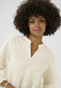 Kreminė palaidinė su V formos iškirpte, puošta sudėtingais gėlės raštais ir ilgomis rankovėmis, pagaminta iš lengvo, tekstūruoto audinio.