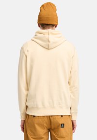 Sudadera de color crema con una textura suave, que cuenta con una capucha con cordón y puños de canalé, combinada con pantalones de trabajo beige.
