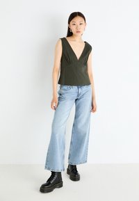 Olive green, sleeveless top with a deep V-neck, gathered detailing, paired with light denim wide-leg jeans and black lace-up boots.