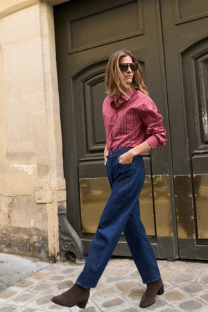 Femme portant des lunettes de soleil, une chemise rayée rouge, un jean bleu et des bottines marron marchant dans une rue pavée près d'une grande porte sombre.