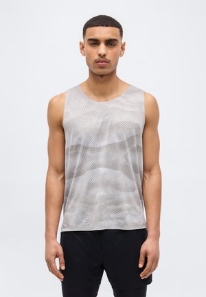 Νεαρός με κοντά μαλλιά που φοράει γκρι αμάνικο τοπ από δίχτυ και μαύρο σορτς, στέκεται μπροστά σε απλό φωτεινό φόντο.