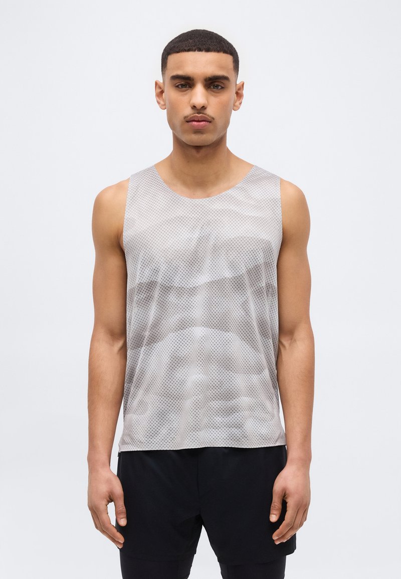 Νεαρός με κοντά μαλλιά που φοράει γκρι αμάνικο τοπ από δίχτυ και μαύρο σορτς, στέκεται μπροστά σε απλό φωτεινό φόντο.