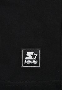 Melns auduma t-krekls ar ribotu tekstūru un kvadrātveida balto etiķeti. Etiķetei ir stilizēts "S" burts un uzraksts "STARTER BLACK LABEL."