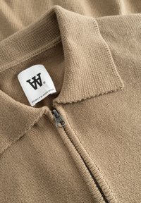 Pull beige en maille avec fermeture éclair, doté d'un large col et d'une fermeture éclair YKK. Étiquette visible avec la marque "Wood Wood".