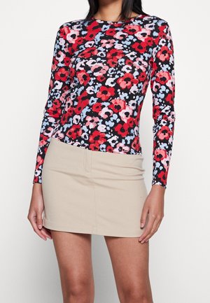 Femme portant un haut à manches longues avec un motif floral rouge, rose et bleu et une mini-jupe beige unie, debout devant un fond blanc.