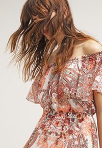 Kobieta z długimi falującymi włosami zasłania twarz, ubrana w sukienkę bez ramiączek w pomarańczowe, czerwone i białe kwiatowe oraz paisley wzory.