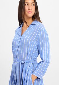 Abito camicia azzurro chiaro a maniche lunghe con strisce bianche verticali, colletto, vita cintata e tasche laterali, realizzato in un tessuto morbido e strutturato.