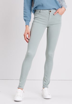 Personne portant un jean skinny bleu clair, un pull bleu et des baskets blanches, avec une main dans la poche et l'autre détendue le long du corps.