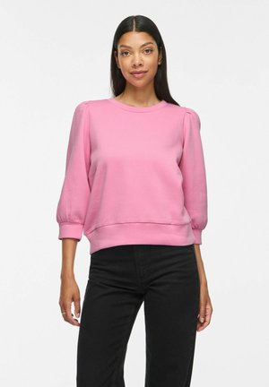 Donna con lunghi capelli neri che indossa un top rosa con maniche a sbuffo e pantaloni neri, in piedi davanti a uno sfondo bianco semplice.